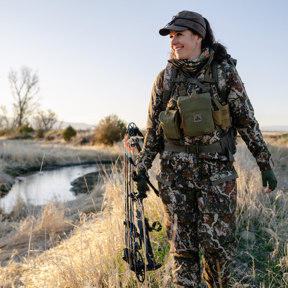 Woman dressed in camouflage with a compound bow in tall grass with a small stream in the background