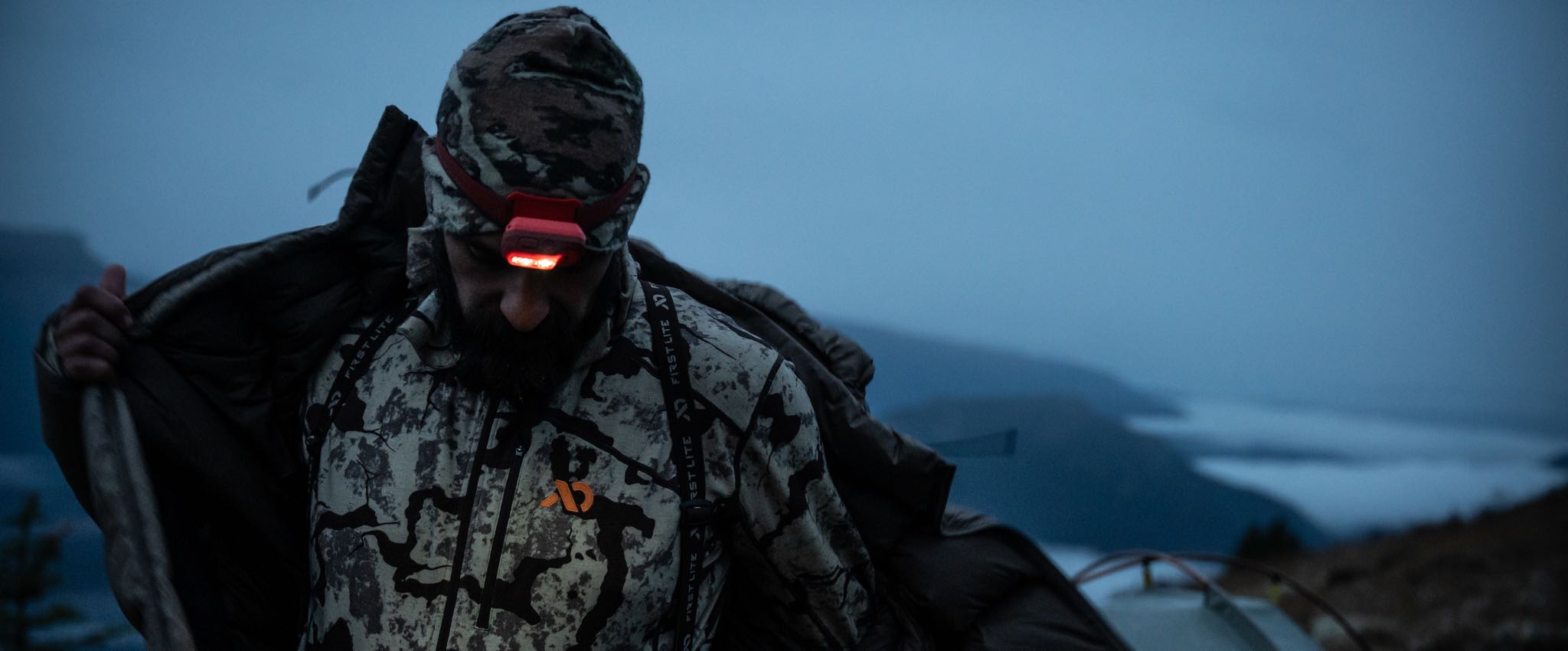 Red headlamp glowing on a person wearing a First Lite camo jacket and hat in low light.